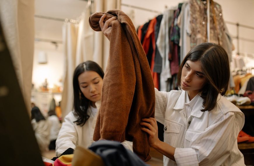 Two women examining a brown coat in a clothing store, surrounded by racks of clothes, indoor lighting