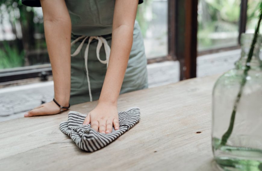 Person wiping wooden table with striped cloth, wearing apron, glass vase on side, natural light background