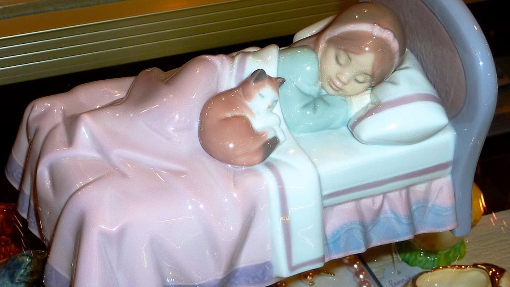 Ceramic figurine of sleeping child with teddy bear in bed, featuring pastel pink and blue colors with glossy finish