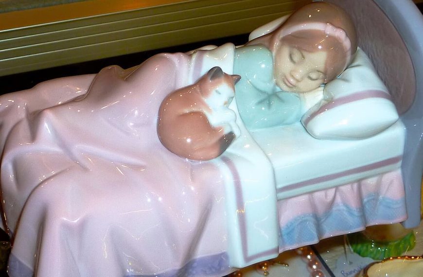 Ceramic figurine of sleeping child with teddy bear in bed, featuring pastel pink and blue colors with glossy finish