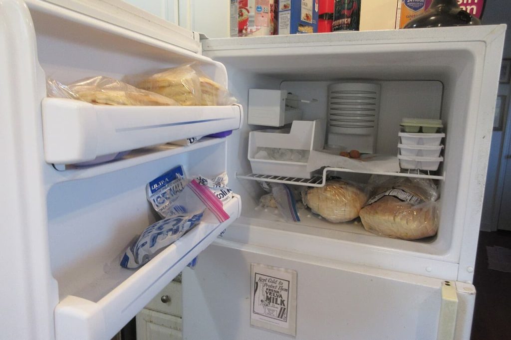 Open refrigerator freezer compartment containing bagged bread, ice trays, frozen items, and ice packs in door shelve