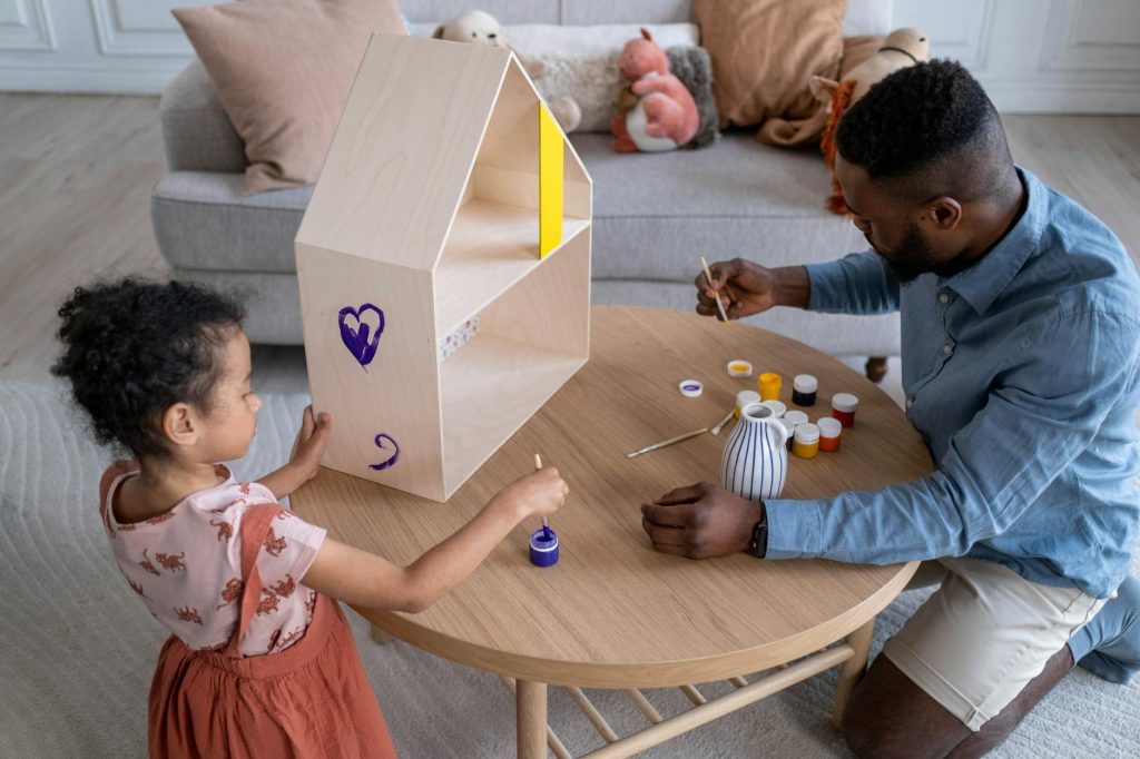 A father and daughter enjoy painting a wooden dollhouse together indoors