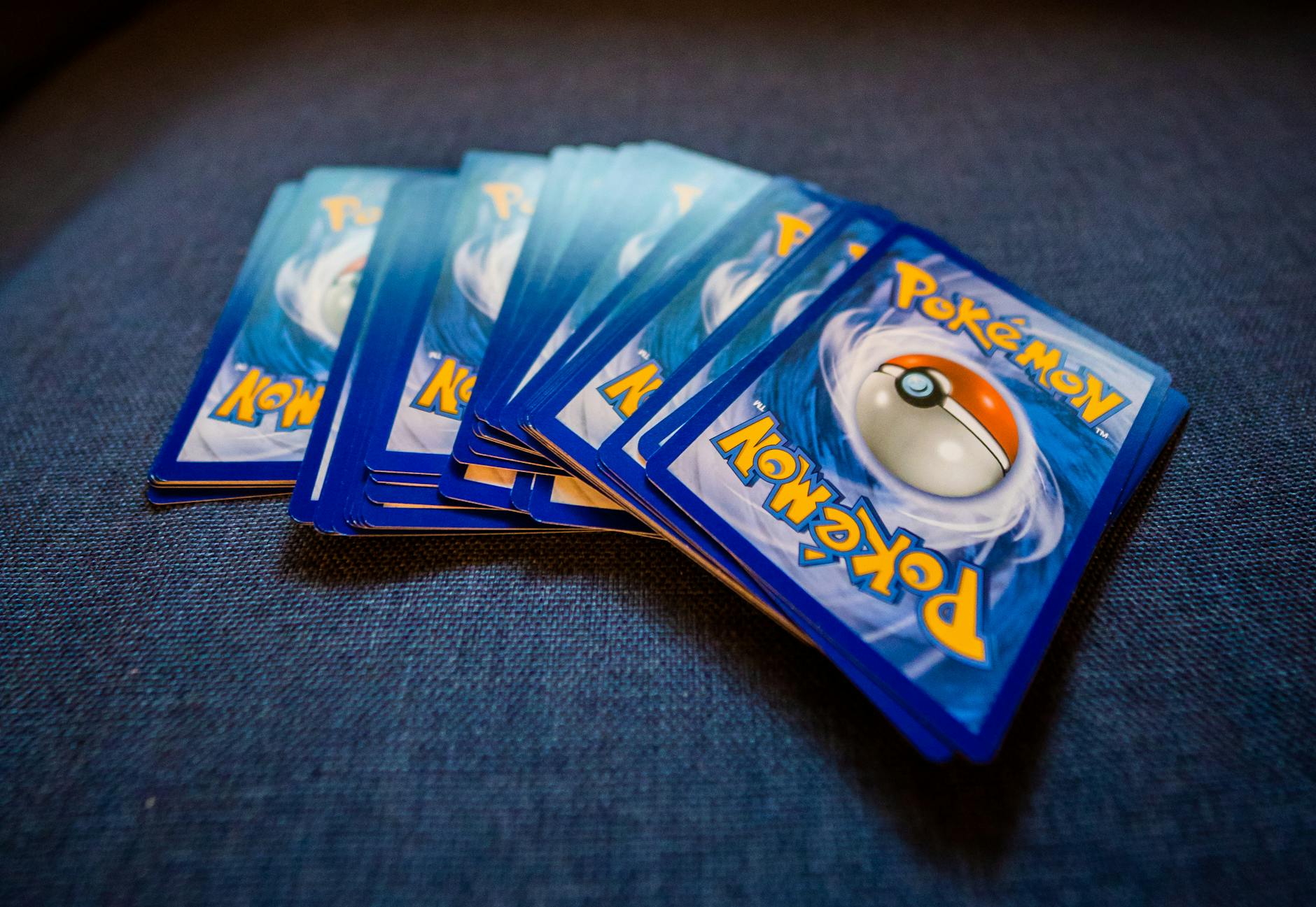 Stack of Pokémon trading cards, blue backs with Poké Ball design, laid out on a dark blue fabric surface
