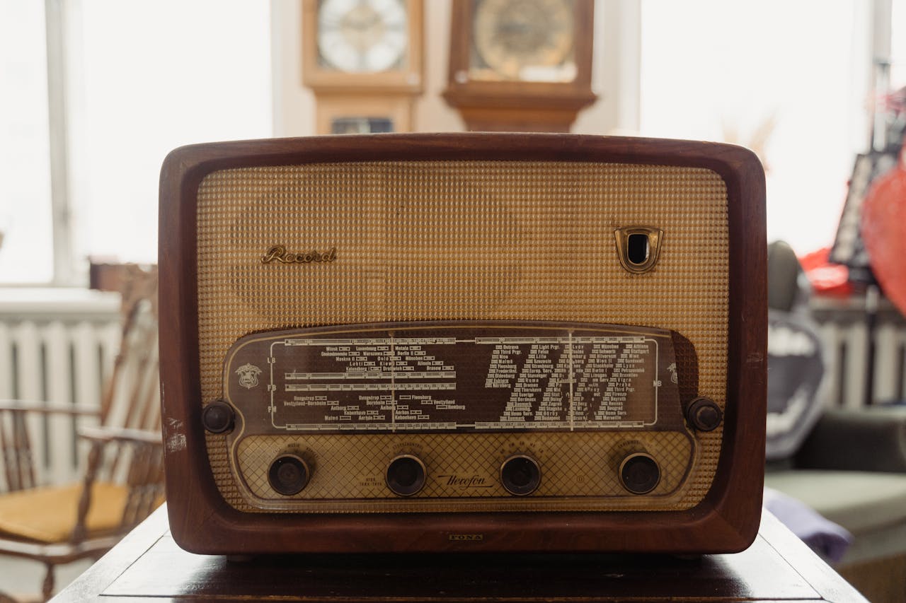 Vintage wooden radio with beige cloth speaker cover. The "Record" brand device features a dark station dial displaying frequency markings, four control knobs along the bottom, and decorative gold emblem. Wall clocks visible in the background