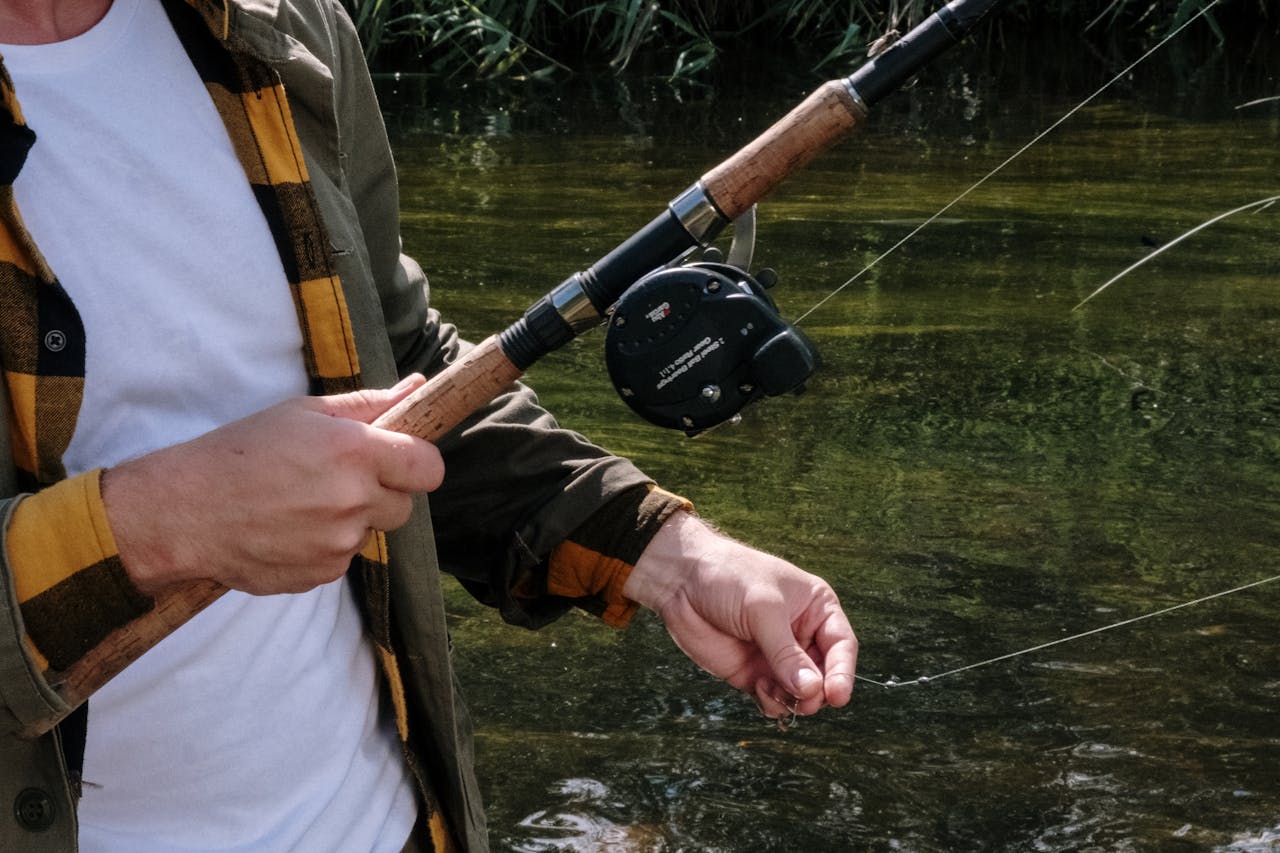 close-up of person wearing yellow plaid shirt holding vintage fishing rod and reel by a calm riverbank.Would you like a second alt text variation for more flexibility