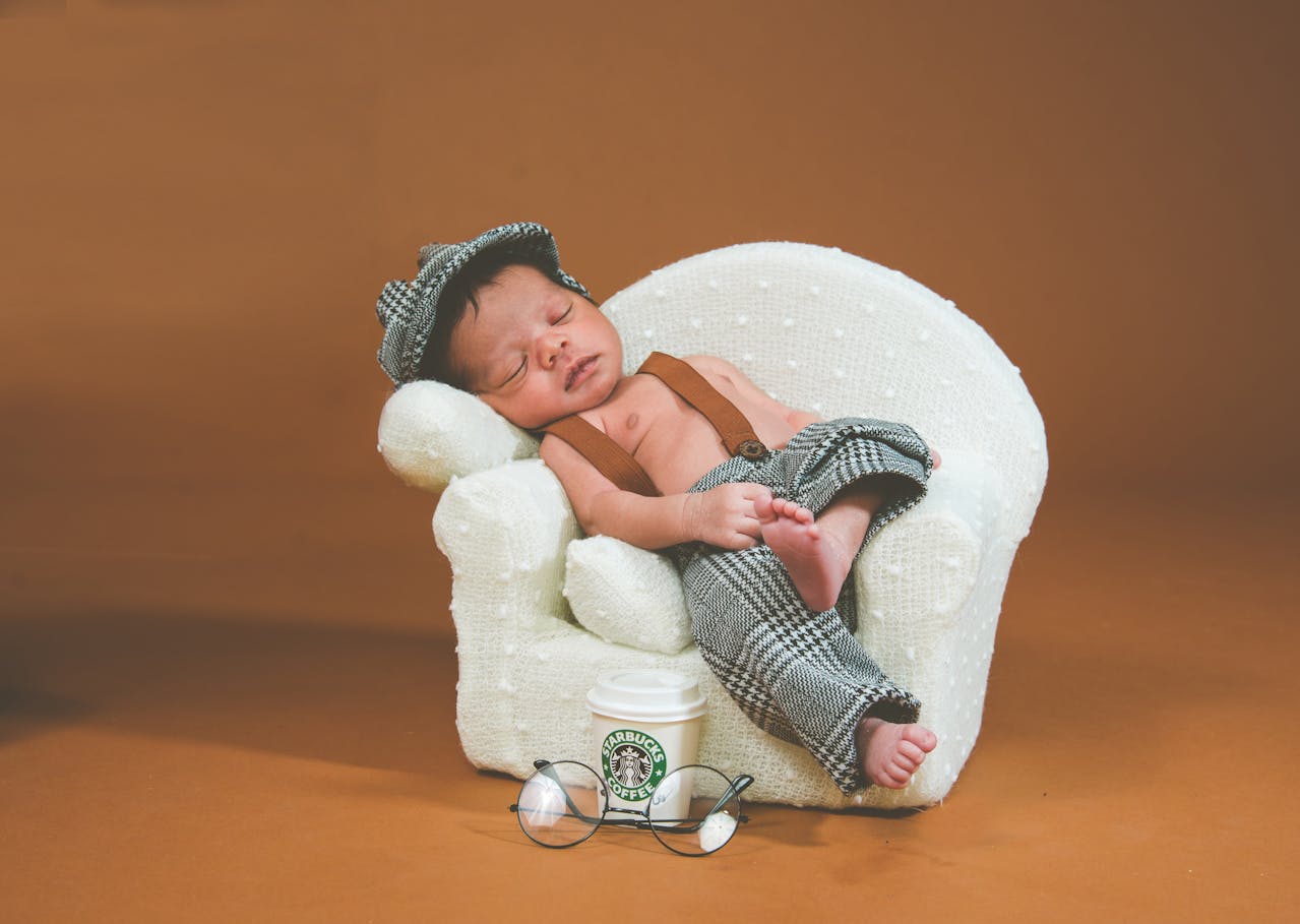 A sleeping newborn baby in a professional photo shoot, wearing a tweed newsboy cap and pants with brown suspenders, reclining on a miniature white armchair. A small Starbucks coffee cup and round glasses are positioned as props in front of the chair