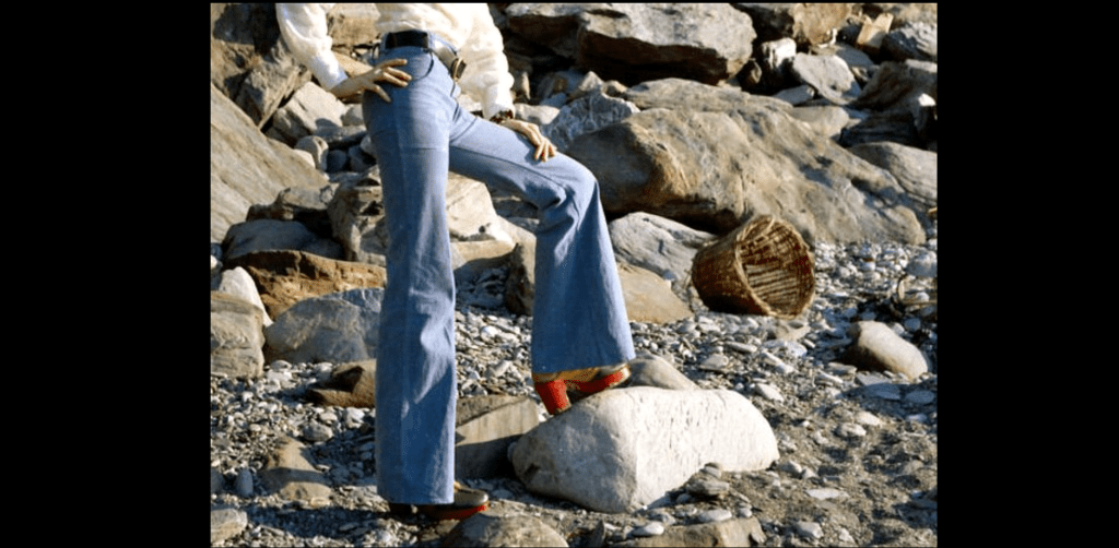 Person wearing high-waisted bell-bottom jeans, standing on rocky beach, red platform shoes, wicker basket in background