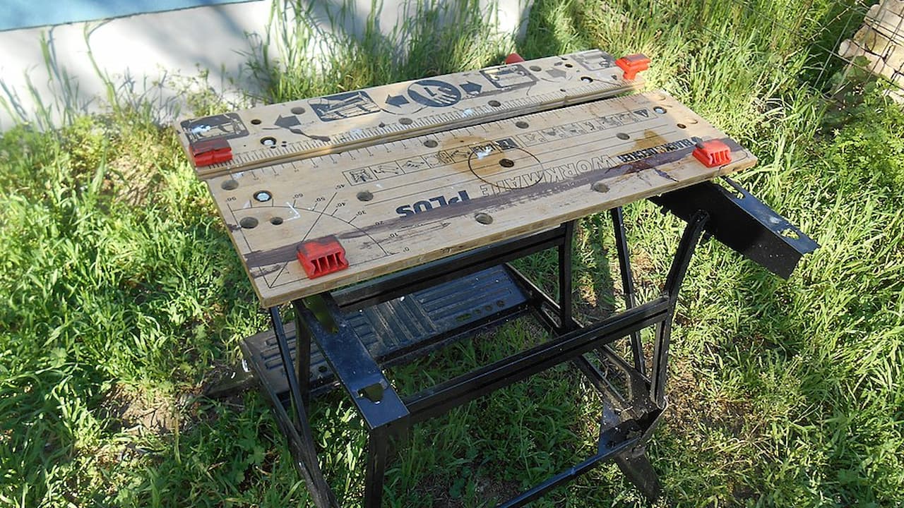 Folding portable workbench, wooden top with printed measurements and angles, red clamps on corners, black metal frame, set on grassy outdoor surface
