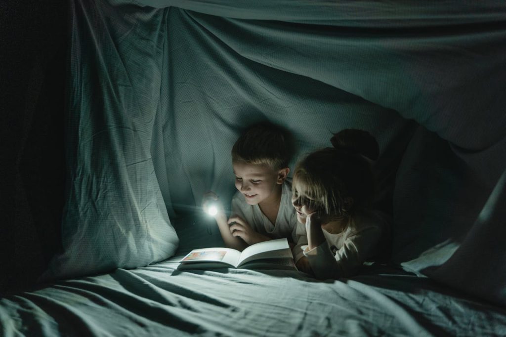 Two children reading a book by flashlight inside a cozy blanket fort