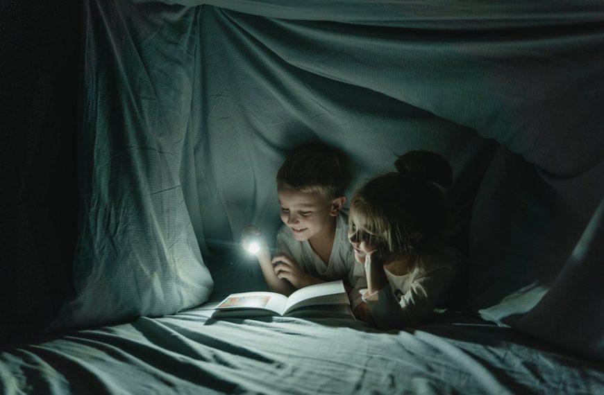 Two children reading a book by flashlight inside a cozy blanket fort