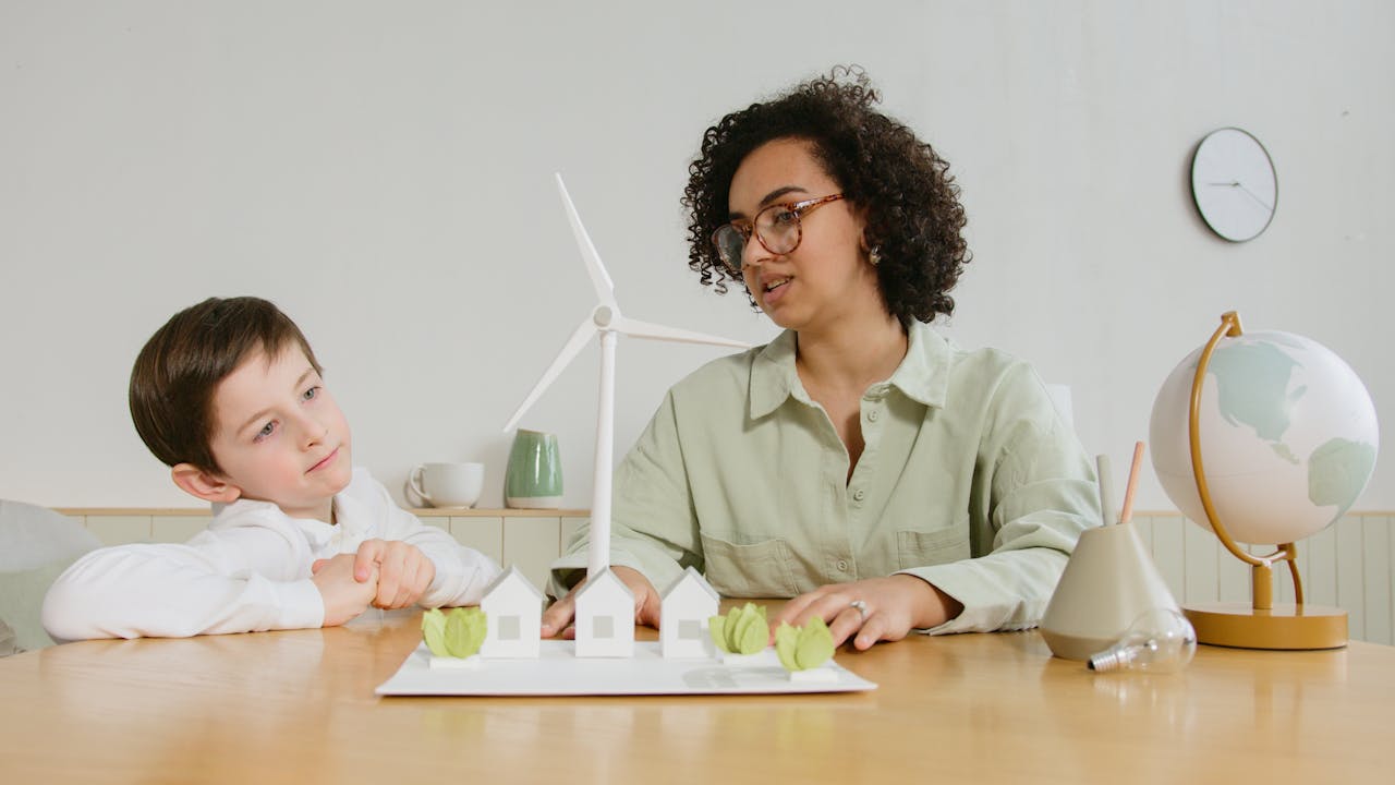 Adult and child sitting at a table, discussing a wind turbine model with miniature houses and trees, educational setting