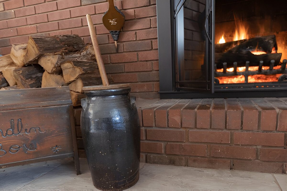 Clay churn with wooden stick, placed near brick fireplace, fire burning warmly in background, stacked firewood on brick shelf, rustic setting, traditional interior décor, cozy and vintage atmosphere