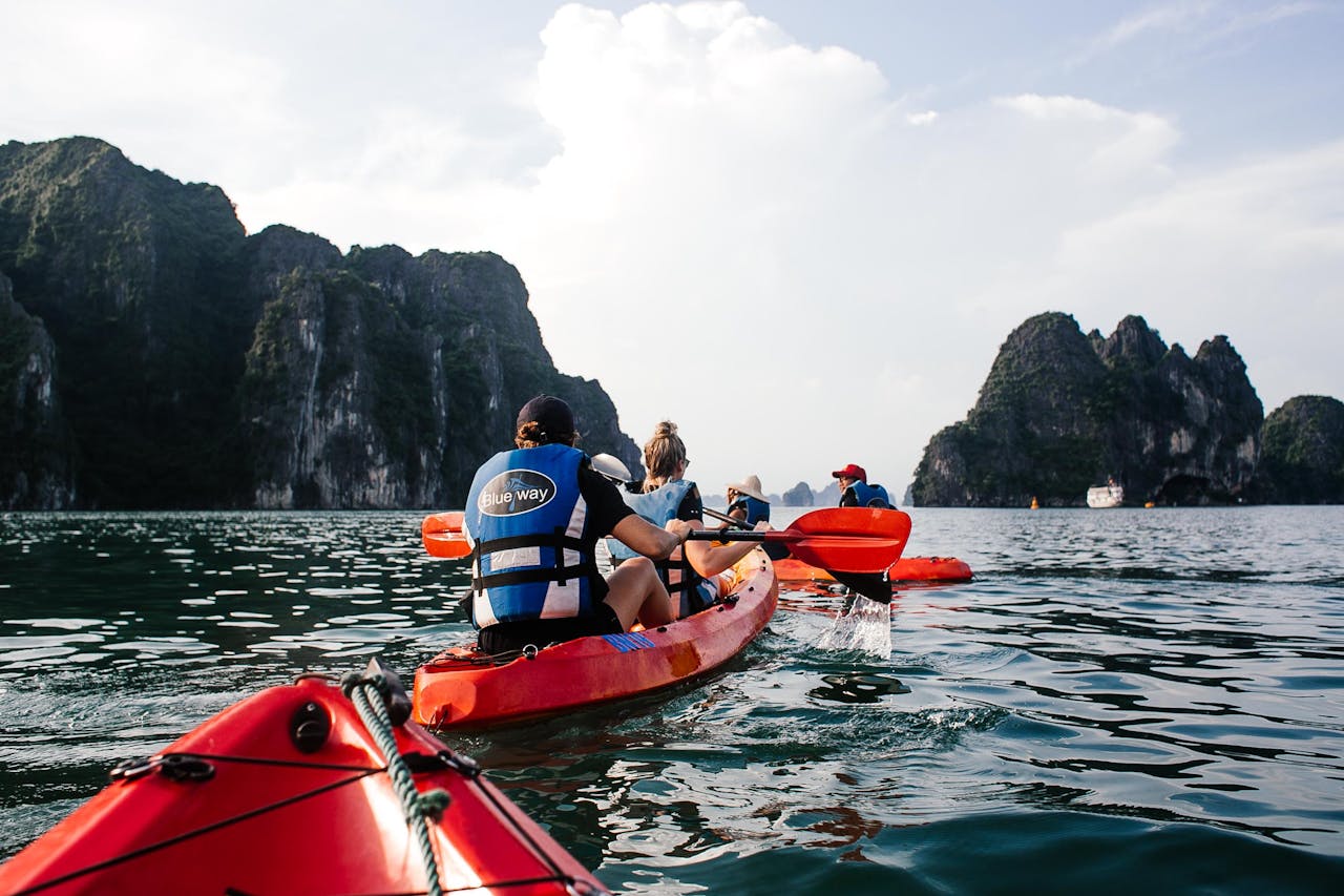 People kayaking on calm water, wearing life jackets, surrounded by tall rocky cliffs, under a clear blue sky