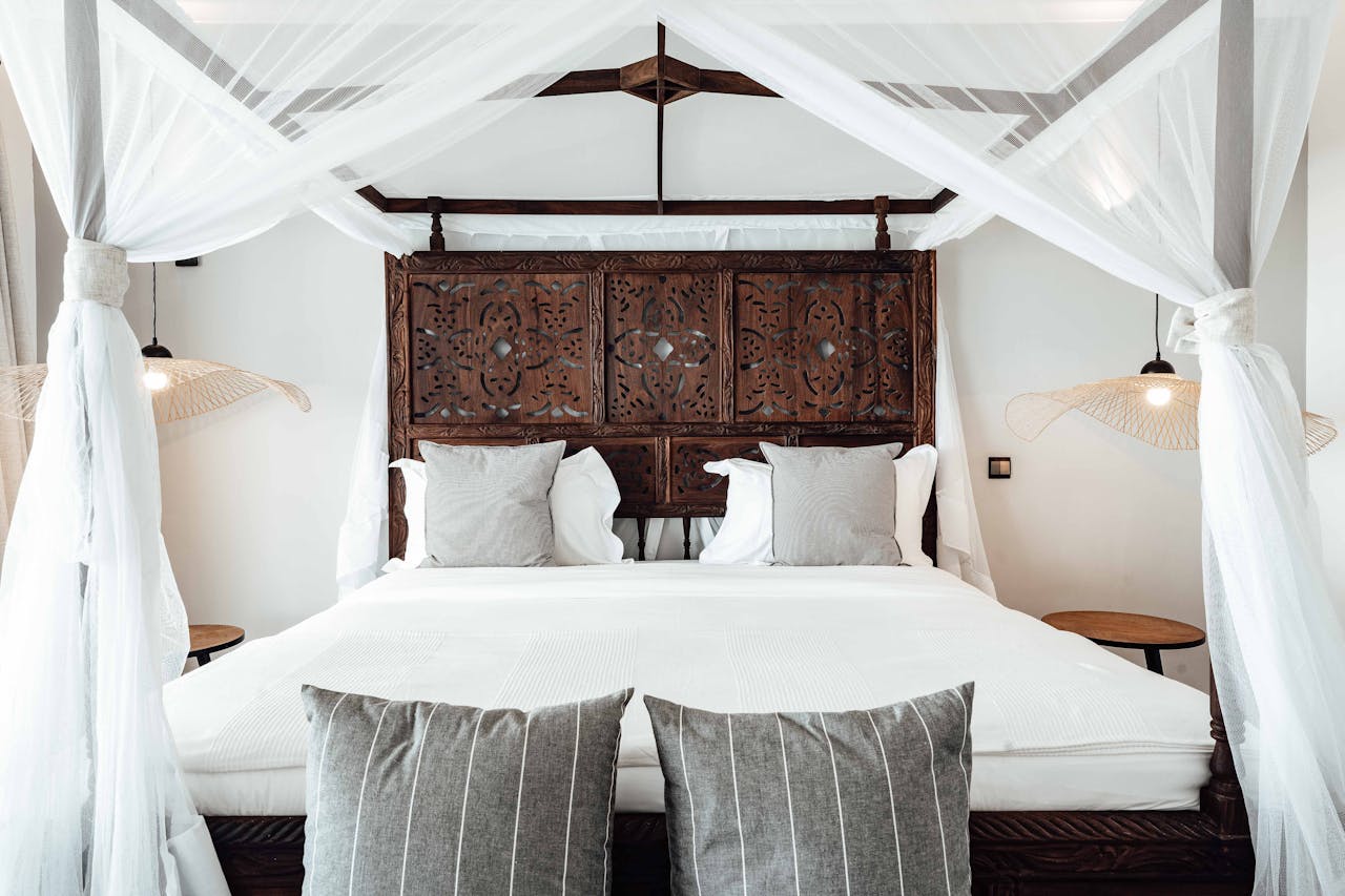 Canopy bed with white curtains, carved wooden headboard, white bedding, grey accent pillows, modern pendant lights, minimalistic bedroom decor