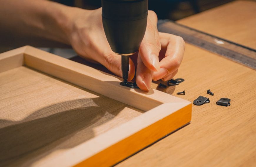 Close-up of a person using a power drill, attaching a black hinge to a wooden frame, small screws and metal brackets scattered on a wooden table