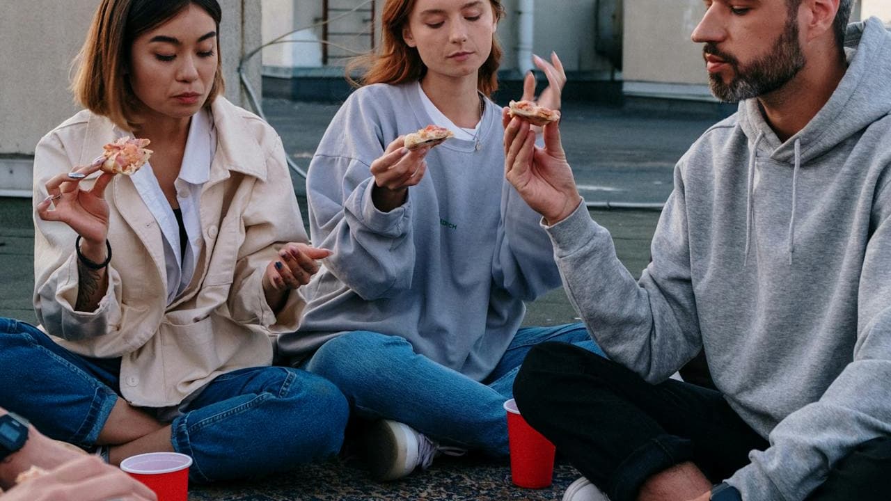 Three people sitting cross-legged on a rooftop, each holding a slice of pizza, wearing casual clothes like hoodies and jackets, red plastic cups placed on the ground