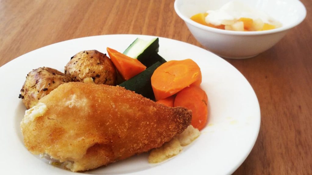 A plate of breaded chicken Kiev served with roasted potatoes, steamed carrots, and zucchini slices sits on a wooden table, accompanied by a bowl of mixed fruit topped with cream in the background