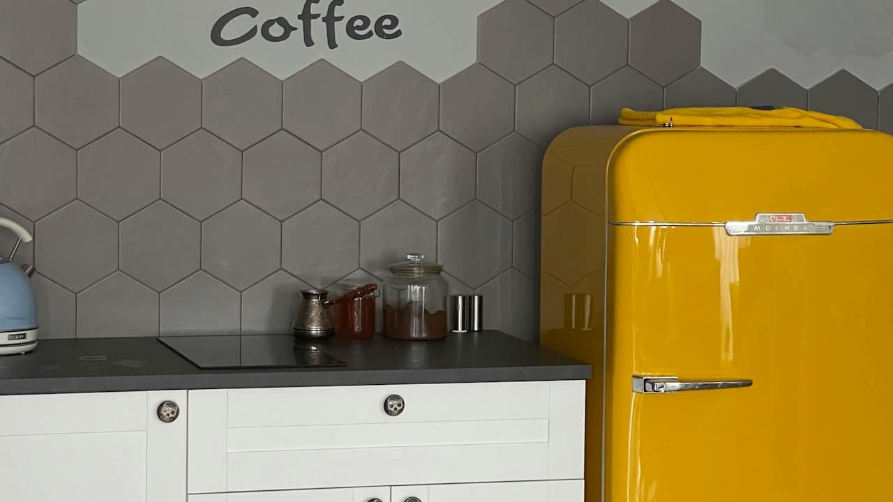 A modern kitchenette features white cabinets, a matte black countertop, a bright yellow retro-style fridge, and a hexagonal grey tile backsplash. A wall decal reads "KEEP CALM AND DRINK Coffee" in bold black text