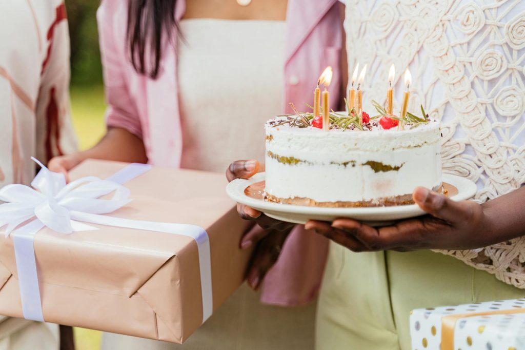 Two people holding a birthday cake with lit candles and a wrapped gift, dressed for an outdoor celebration