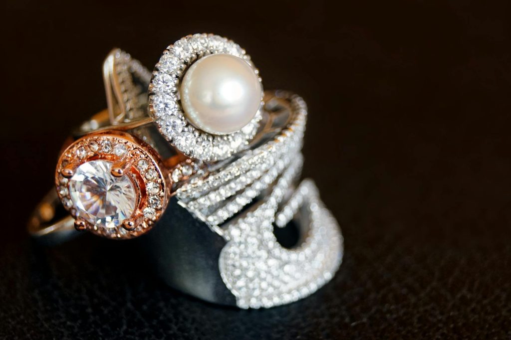 Three elegant rings displayed on a black surface, one featuring a large white pearl surrounded by diamonds, another with a round white gemstone encircled by rose gold and diamonds, and the third with a modern, abstract diamond-encrusted