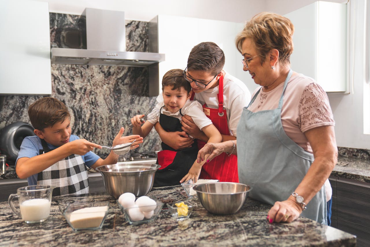 Family in kitchen baking together, young boy sifting flour, child held by adult with whisk, grandmother smiling, ingredients on counter