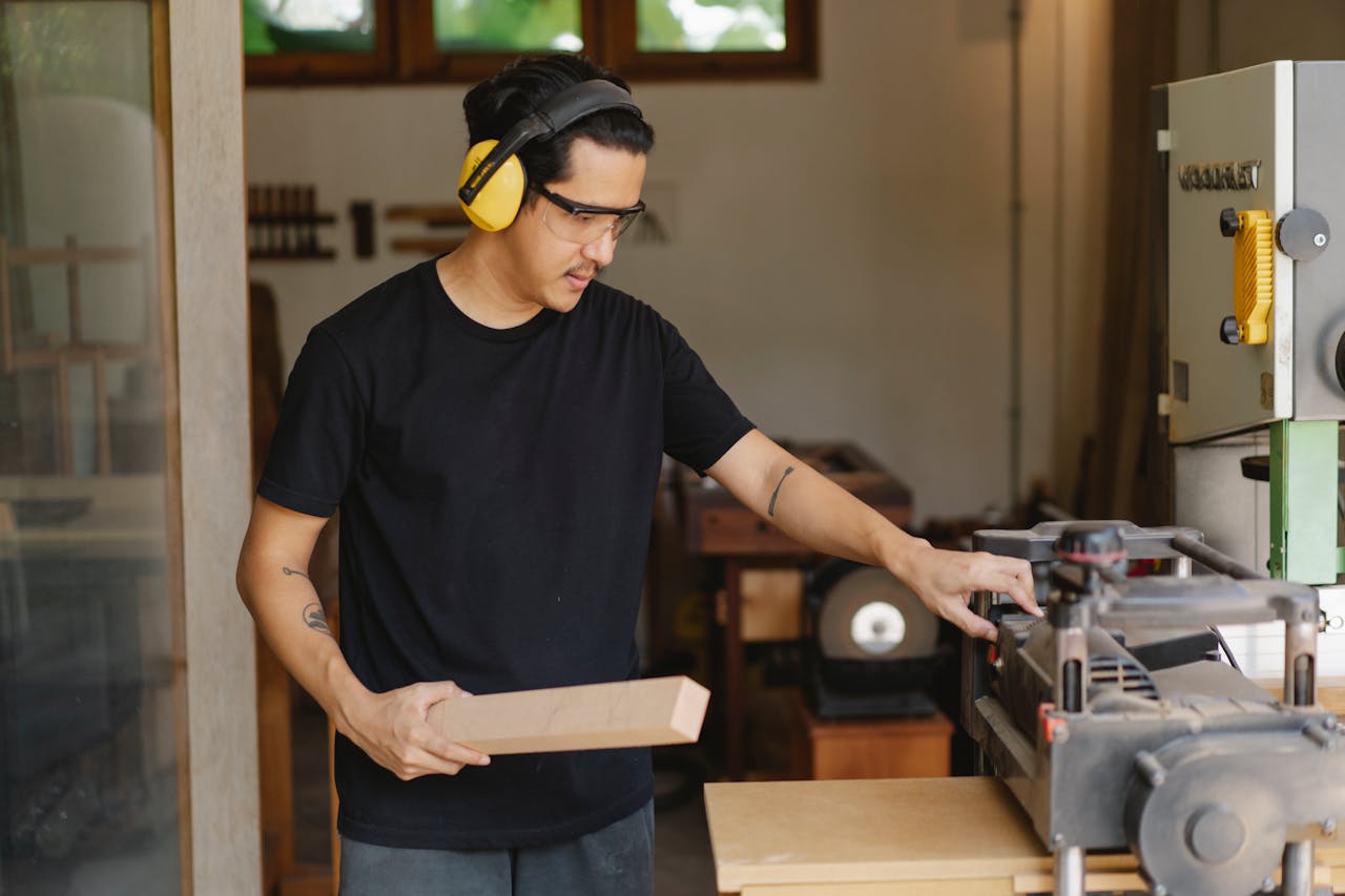 Man wearing safety glasses and yellow ear protection, operating a Dewalt planer, holding a wooden board, standing in a woodworking workshop