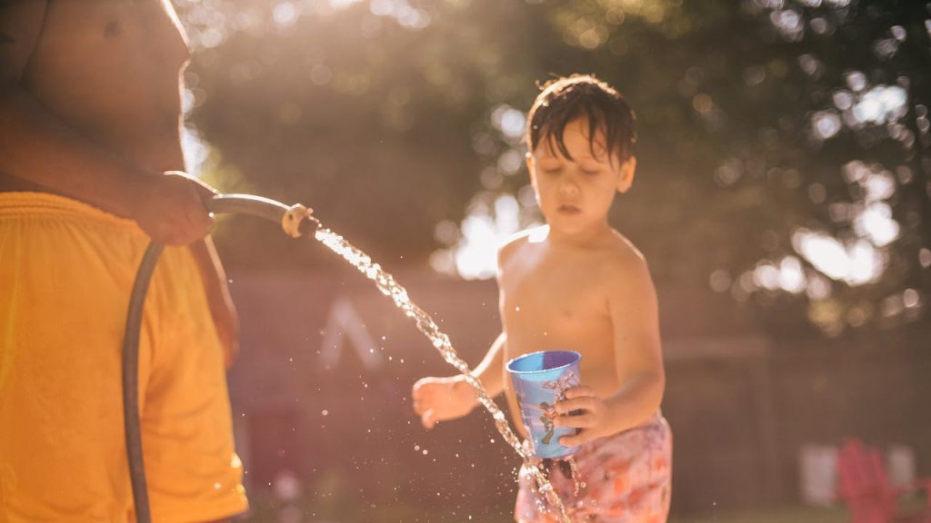 child in swim trunks holding blue cup, adult spraying water from garden hose, backyard, sunlit scene, summer playtime