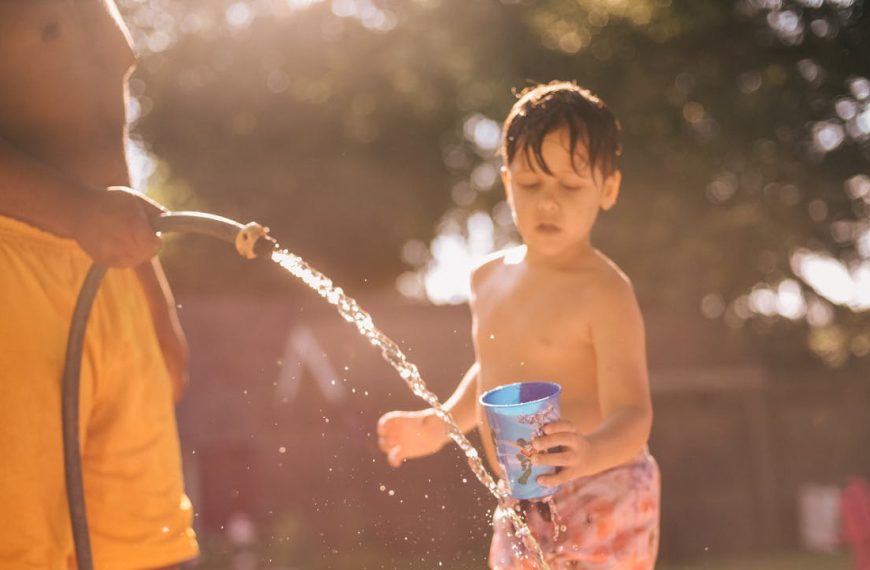 child in swim trunks holding blue cup, adult spraying water from garden hose, backyard, sunlit scene, summer playtime