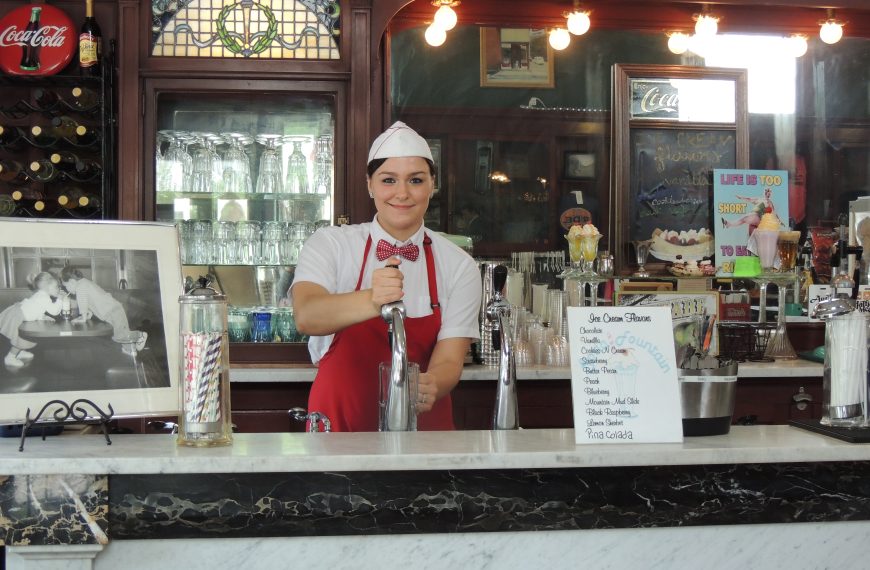 woman in red apron and bow tie, standing behind vintage soda fountain counter, surrounded by milkshake equipment, glassware, retro signage