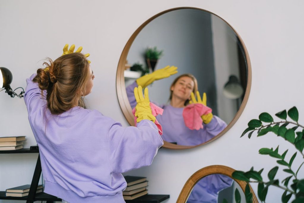 Woman wearing yellow gloves cleaning a round mirror, holding a pink cloth, reflection visible, dressed in a lavender sweatshirt