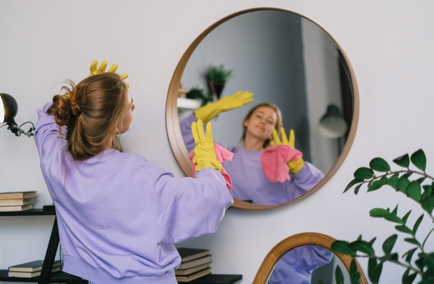 Woman wearing yellow gloves cleaning a round mirror, holding a pink cloth, reflection visible, dressed in a lavender sweatshirt