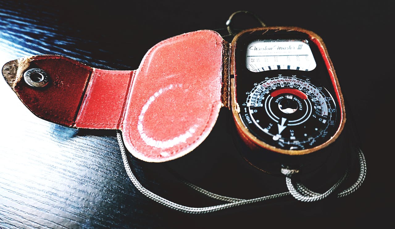 A vintage light meter named "Weston Master III" in an open red leather case. The circular exposure meter shows measurement dials and a needle display, with a thin cord attached to the device