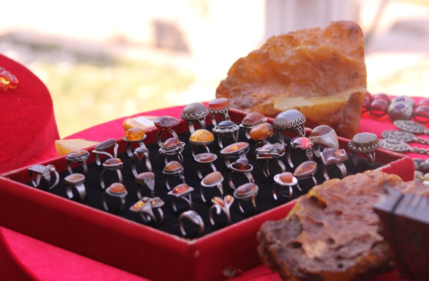 Display of assorted vintage amber rings in a red velvet tray, polished gemstones in silver settings, large amber stones in background
