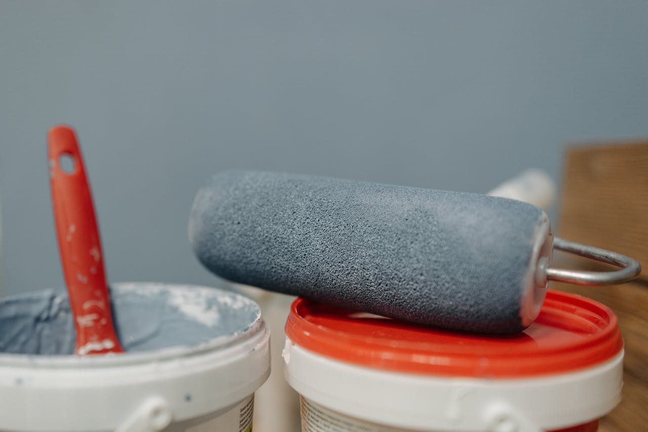 Blue paint roller, red-handled brush in white bucket, open paint containers, soft blue wall background