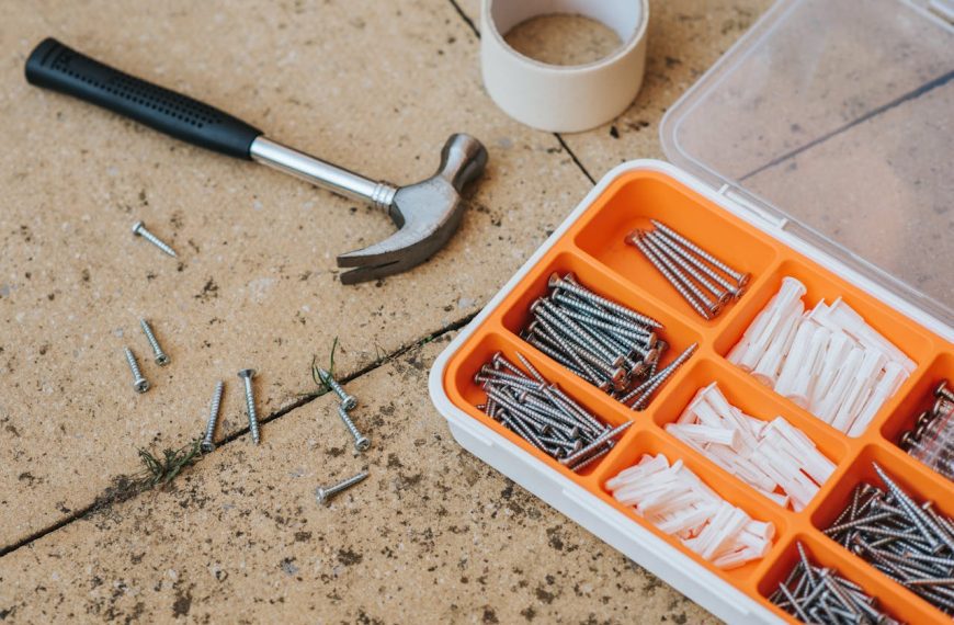 law hammer with black handle lying on stone surface, plastic organizer box filled with assorted screws and wall plugs, scattered screws on ground