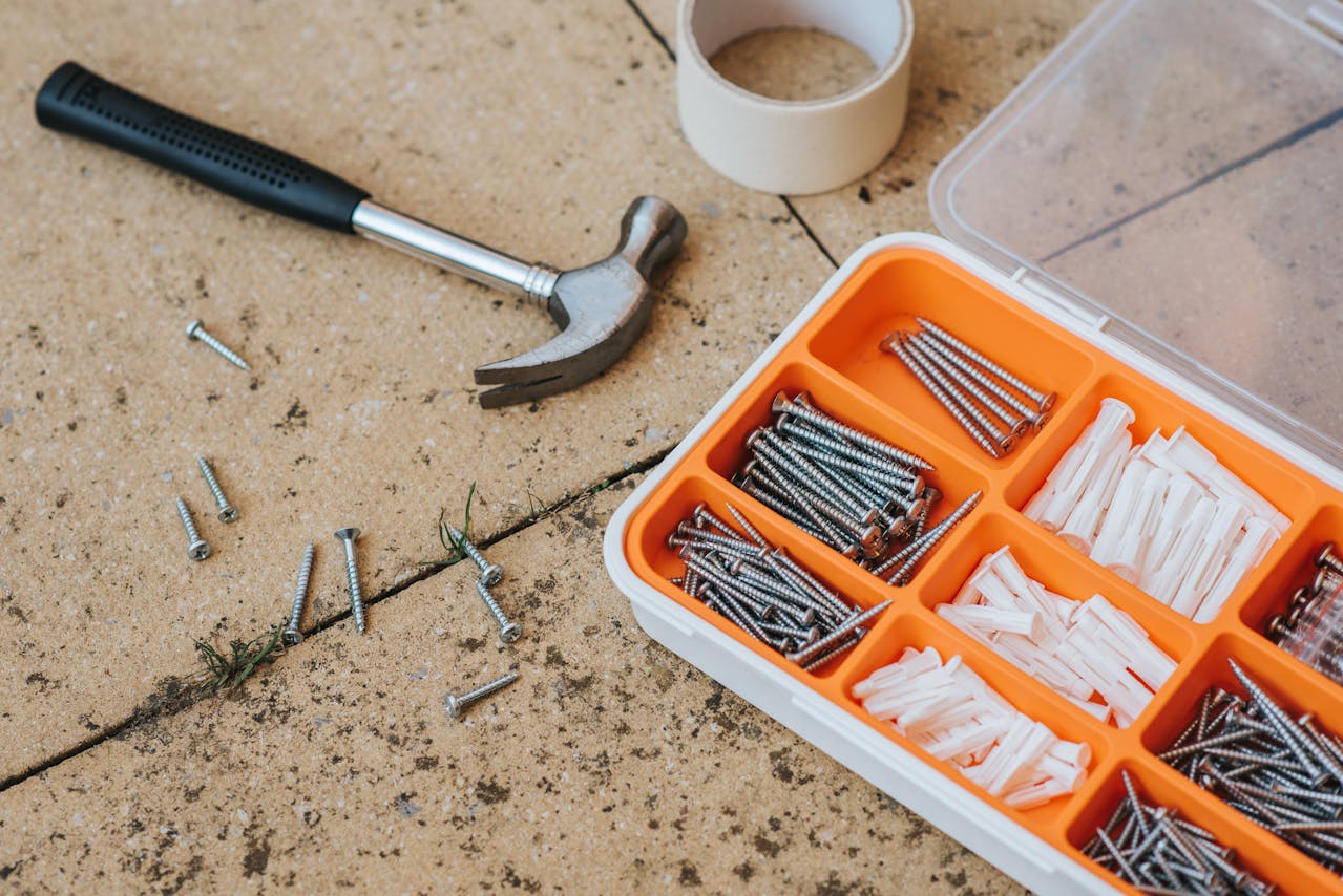 law hammer with black handle lying on stone surface, plastic organizer box filled with assorted screws and wall plugs, scattered screws on ground