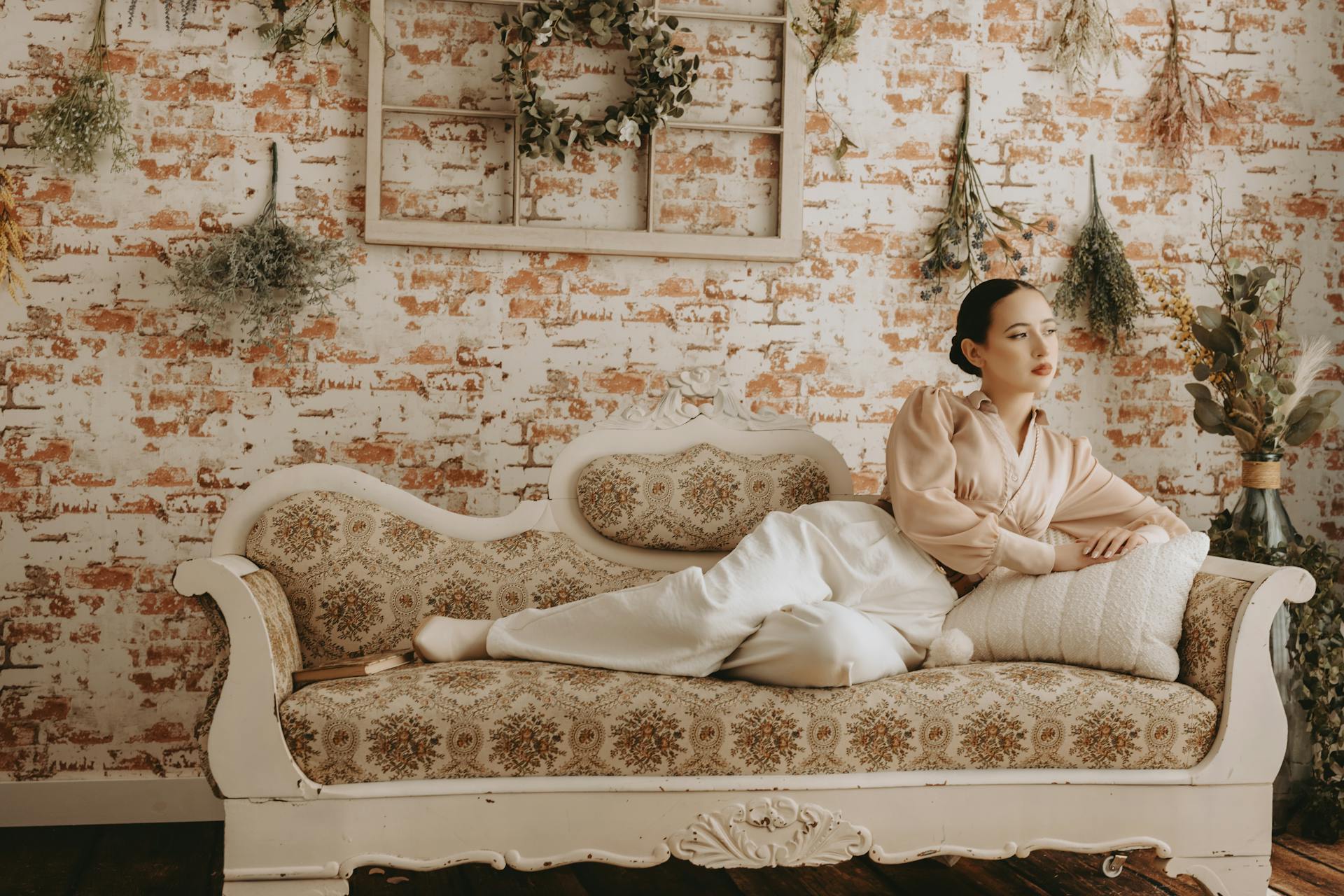 Woman relaxing on a vintage cream-colored couch, floral wallpaper background, soft lighting, dried floral wall décor