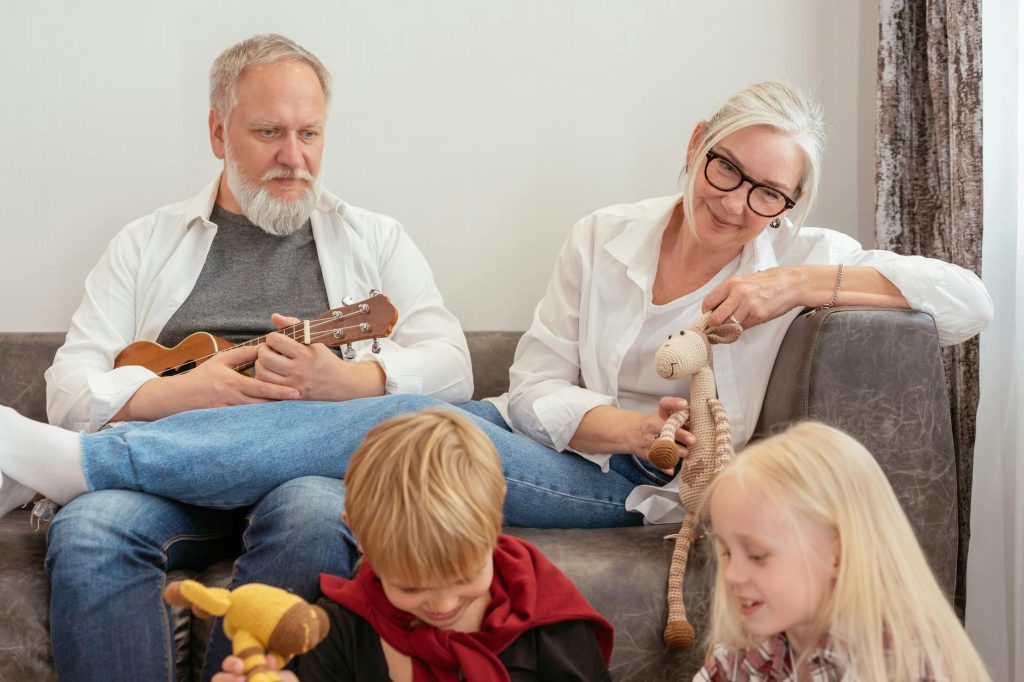 Elderly couple sitting on a couch smiling while two young children play with toys in front of them