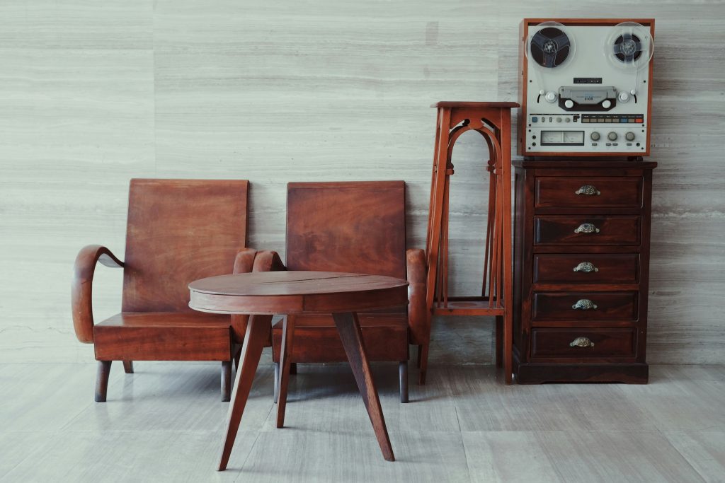 Vintage wooden furniture set, including a mid-century style table, chairs, and a tall chest of drawers, arranged against a neutral-toned wall with tiled flooring