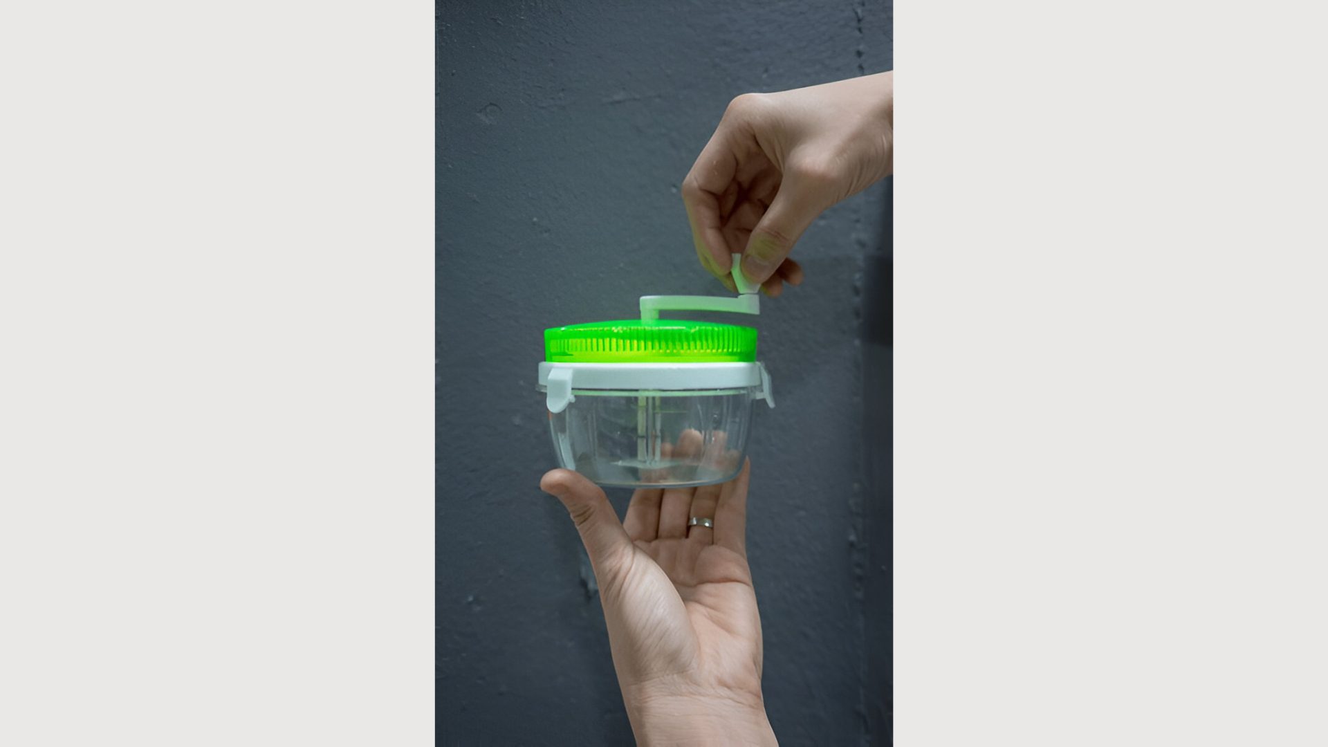 Hand holding empty plastic container of mini kitchen tools for manual food chopper with green lid against grey wall.