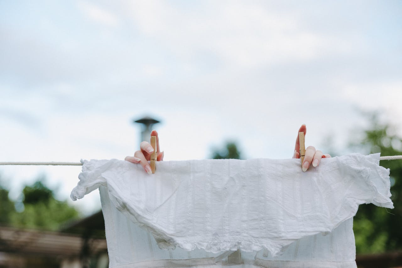 White garment being pinned to a clothesline, two hands holding clothespins, clear sky in background, outdoor setting