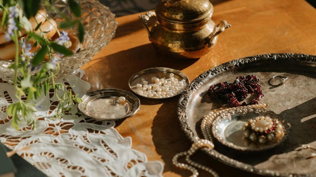 Vintage jewelry and pearls displayed on ornate silver trays, antique brass container in background, soft sunlight casting warm shadows