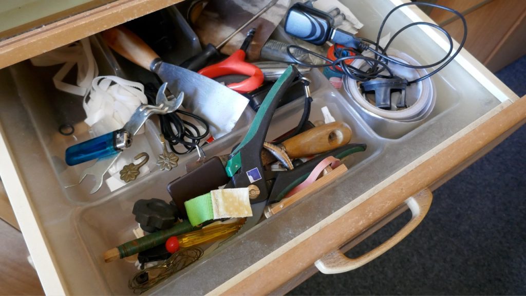 Cluttered wooden toolbox or drawer containing various tools including pliers, screwdrivers, and other hardware items haphazardly stored together. A blue checkmark appears in the corner