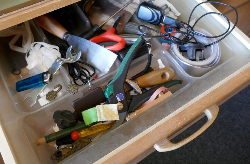 Cluttered wooden toolbox or drawer containing various tools including pliers, screwdrivers, and other hardware items haphazardly stored together. A blue checkmark appears in the corner
