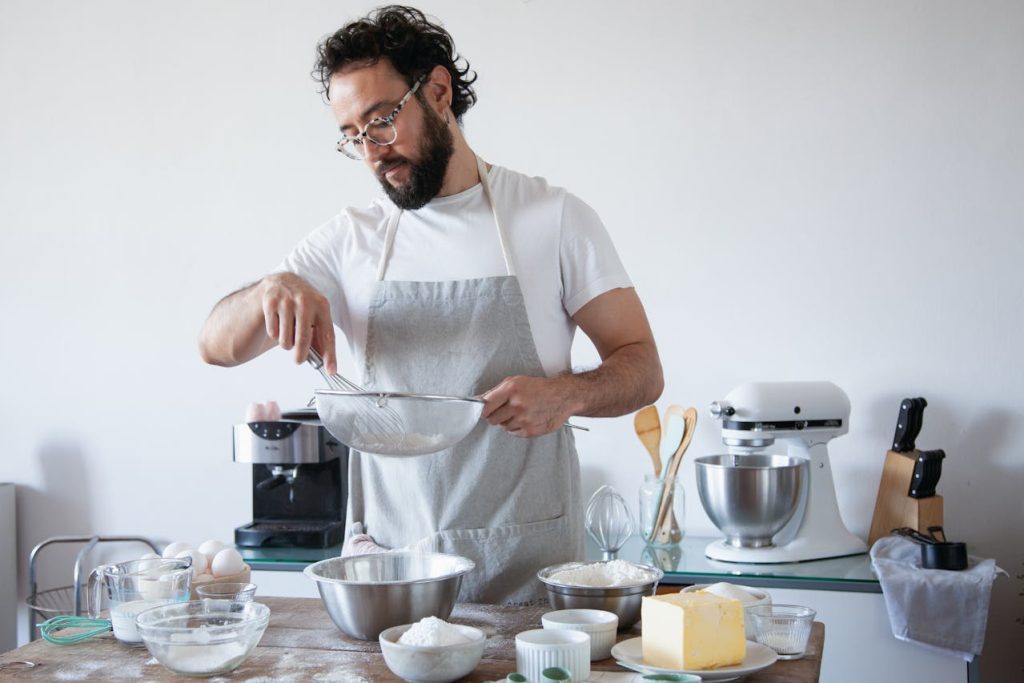 Man wearing glasses and a grey apron, sifting flour into a mixing bowl, surrounded by baking ingredients like butter, sugar, and eggs, standing in a modern kitchen with a stand mixer, utensils