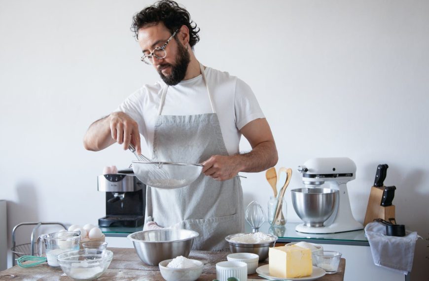 Man wearing glasses and a grey apron, sifting flour into a mixing bowl, surrounded by baking ingredients like butter, sugar, and eggs, standing in a modern kitchen with a stand mixer, utensils