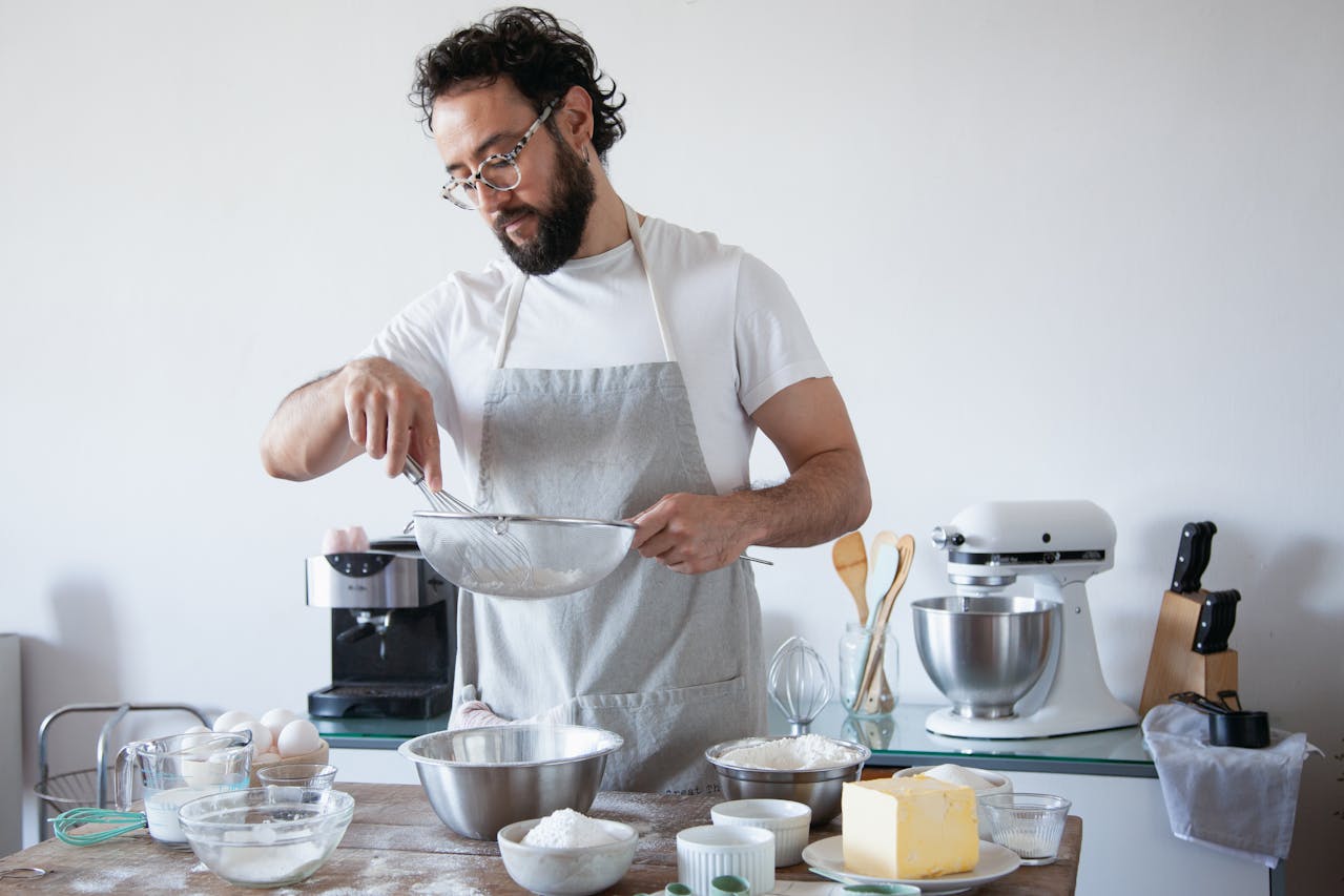 Man wearing glasses and a grey apron, sifting flour into a mixing bowl, surrounded by baking ingredients like butter, sugar, and eggs, standing in a modern kitchen with a stand mixer, utensils
