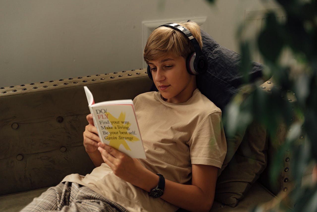 teen boy reading on sofa, wearing headphones, holding inspirational book, calm home setting, relaxing afternoon, quiet solo activity