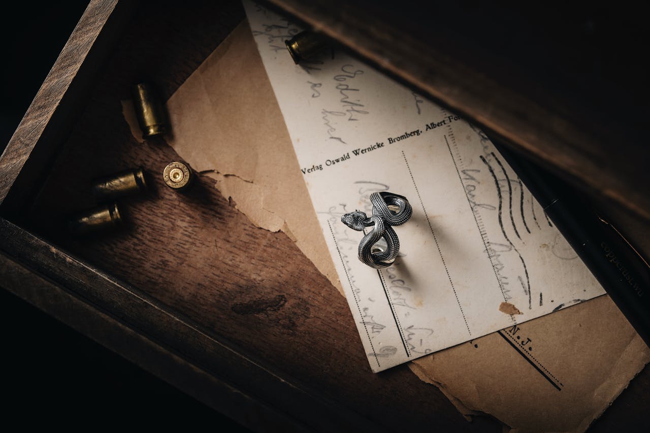 Inside an open wooden drawer: several brass bullet casings on the left, a vintage document with handwriting and printed text in the center, and a small silver snake-shaped pin or brooch resting on the paper