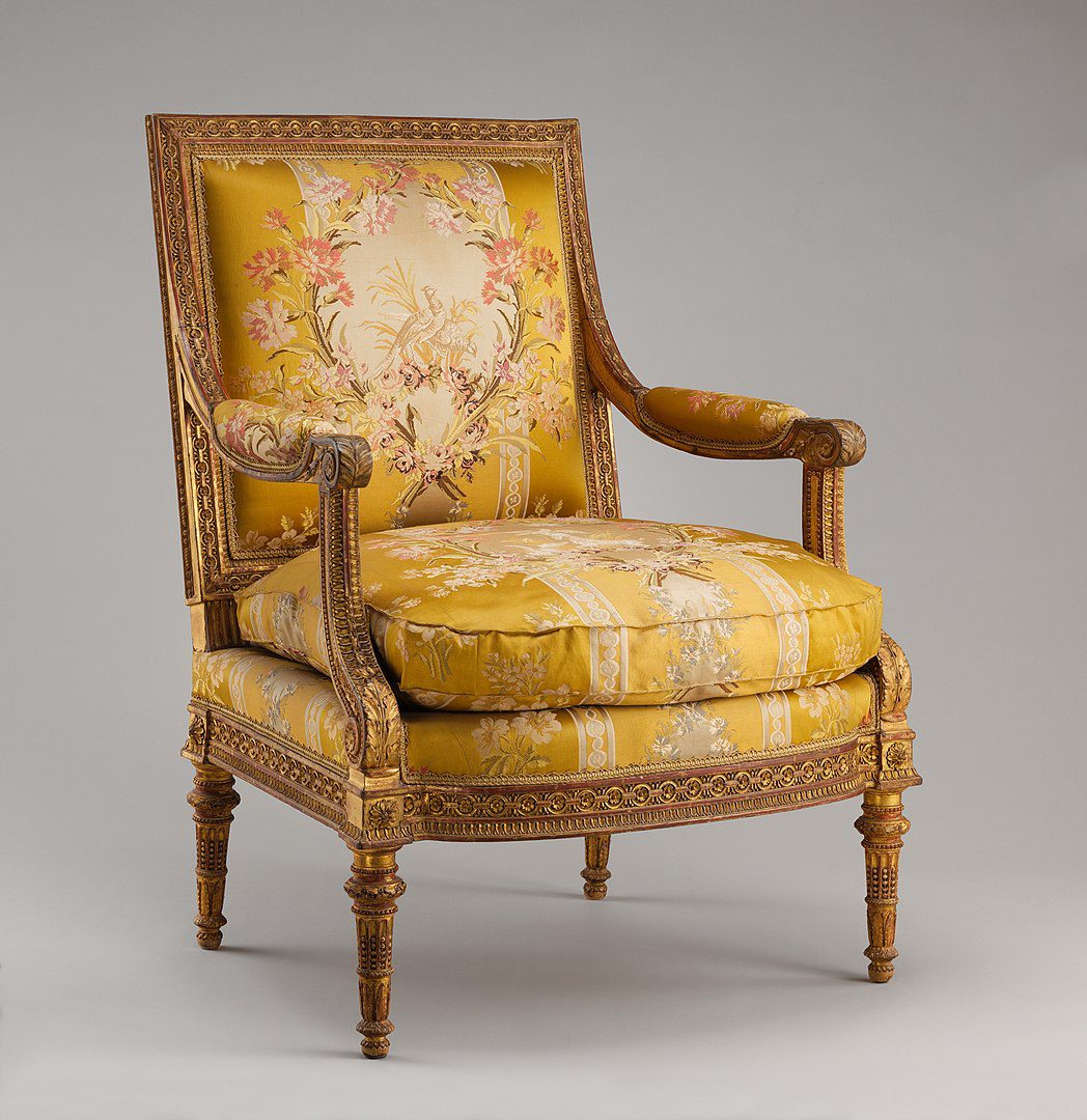 Ornate antique French armchair with carved wooden frame and gilded details. Upholstered in golden yellow silk fabric featuring floral patterns and bird motifs. Louis XVI style with curved arms and decorative scrollwork against neutral background